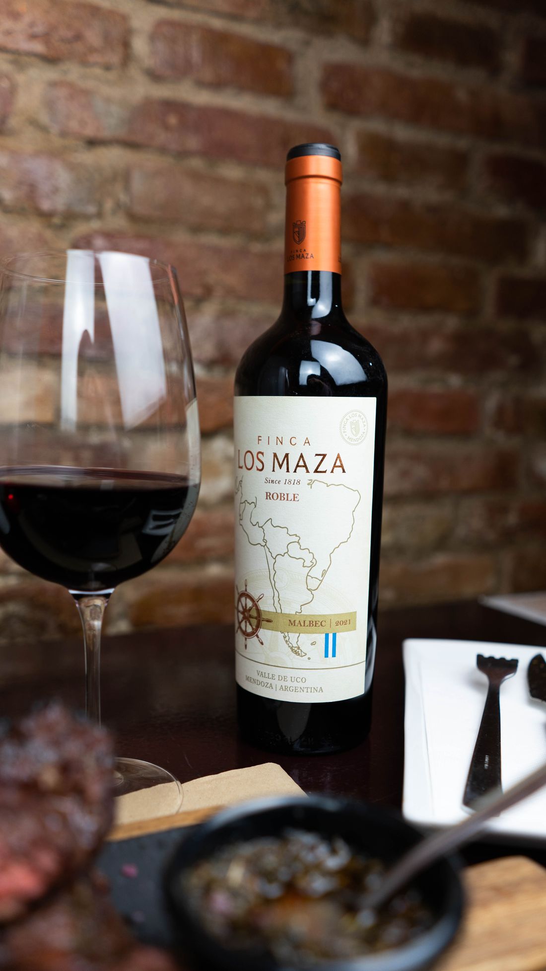 Copa de vino argentino y botella de Malbec sobre una mesa de madera junto a un plato de parrilla